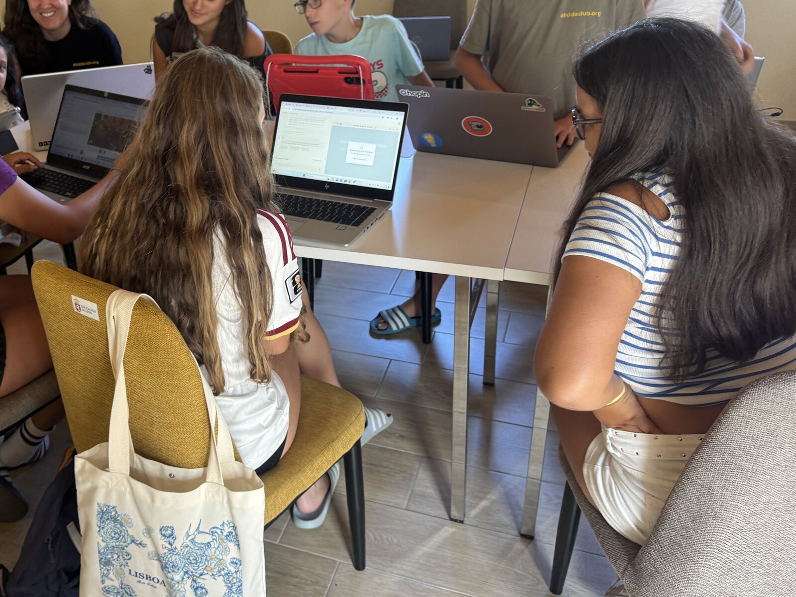 AI Kids Club students working on their projects on laptops around a table, with the instructors alongside them