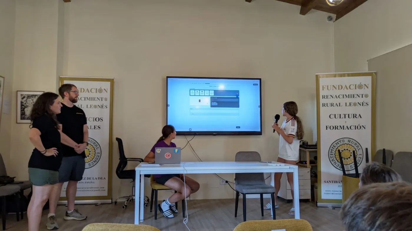 A girl presents with a microphone in front of the big screen, her project open on it, while a teammate works the laptop