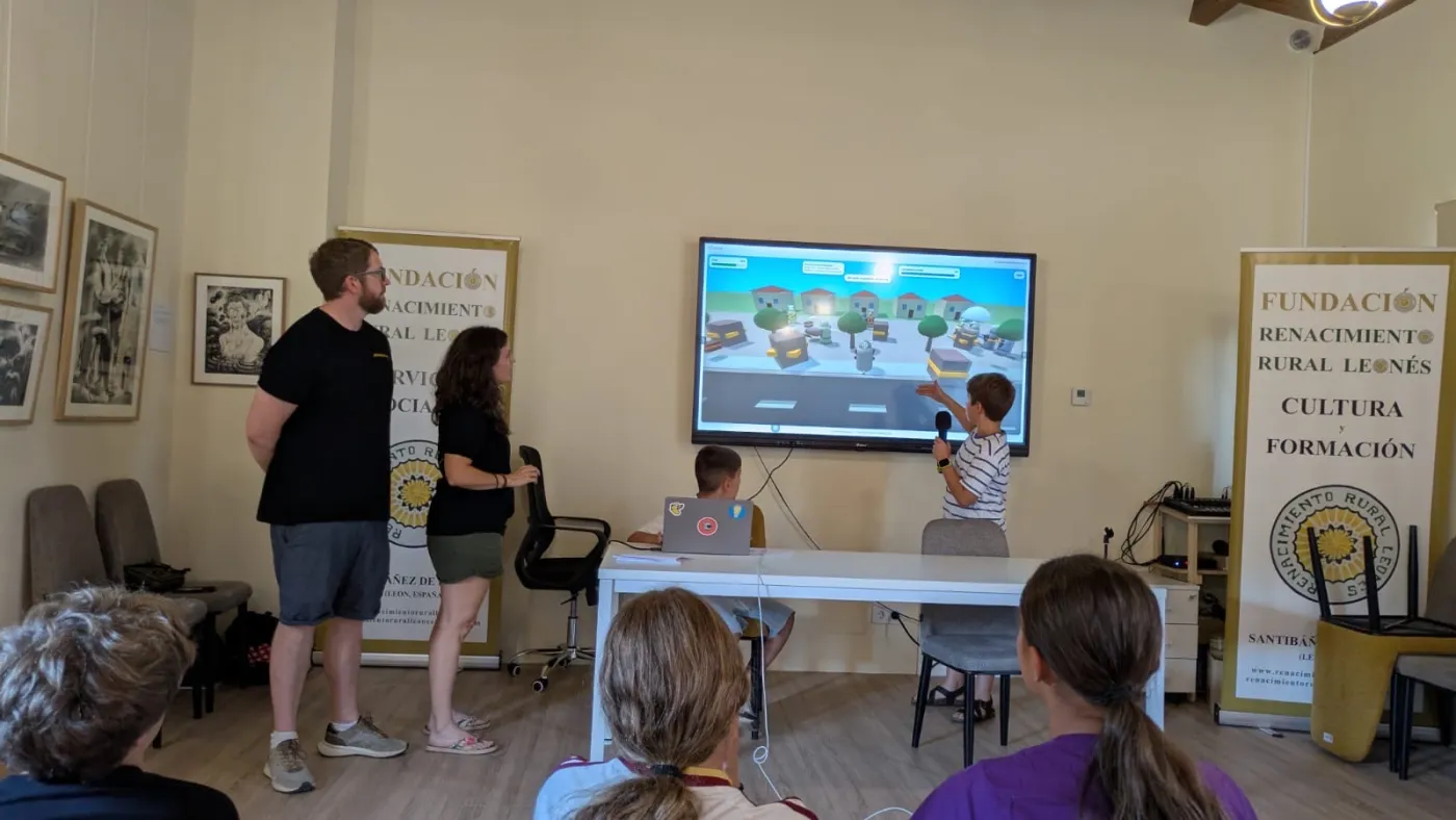 A boy points at his game on the screen as he explains it into the microphone, the audience seated with their backs to the camera in the foreground